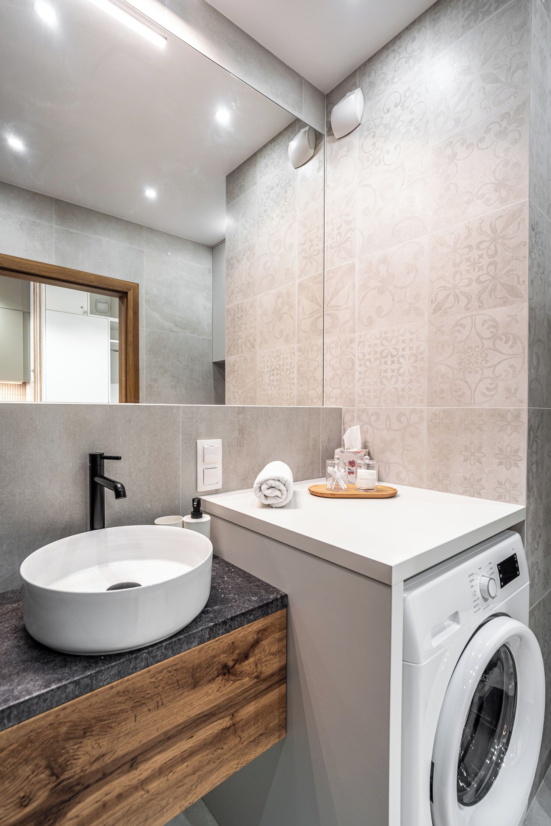 A modern bathroom with a sink, mirror, wall-mounted towel dispenser, and a front-loading washing machine.