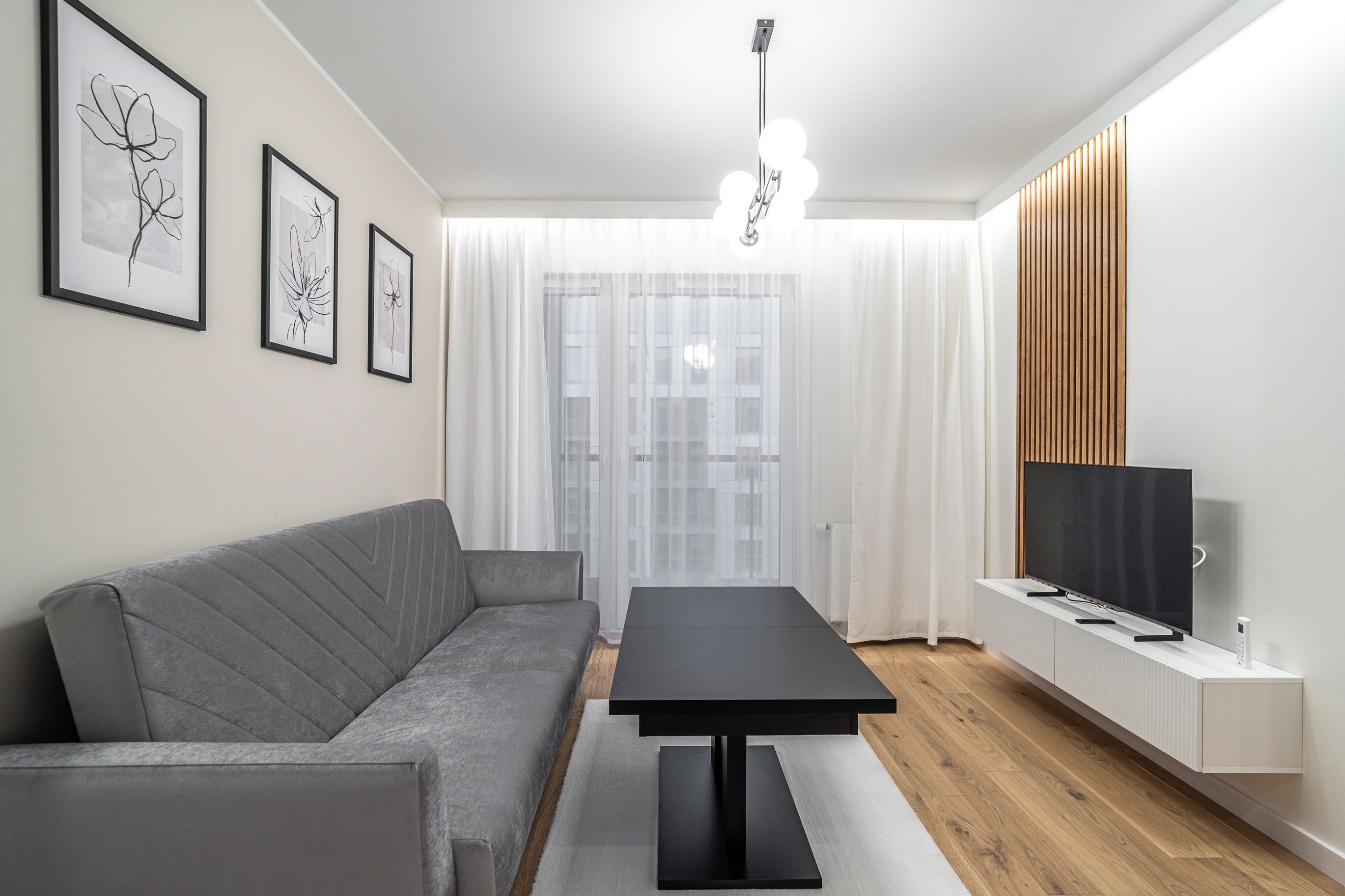 A modern living room with a grey sofa, a black coffee table, a TV on a white cabinet, and framed artwork on the wall.