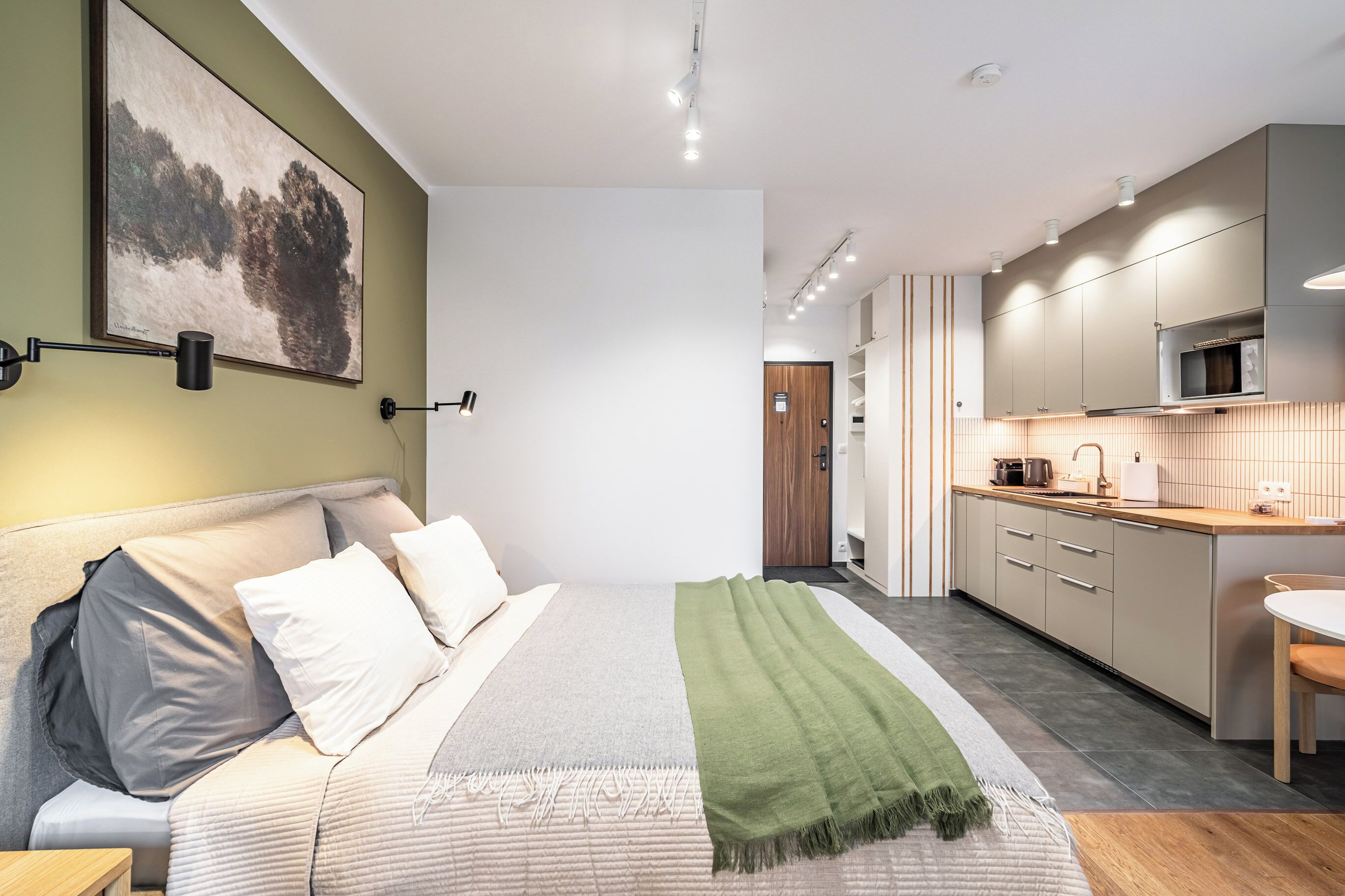 Een moderne hotelkamer met een bed, een groene plaids, een schilderij en een kitchenette.