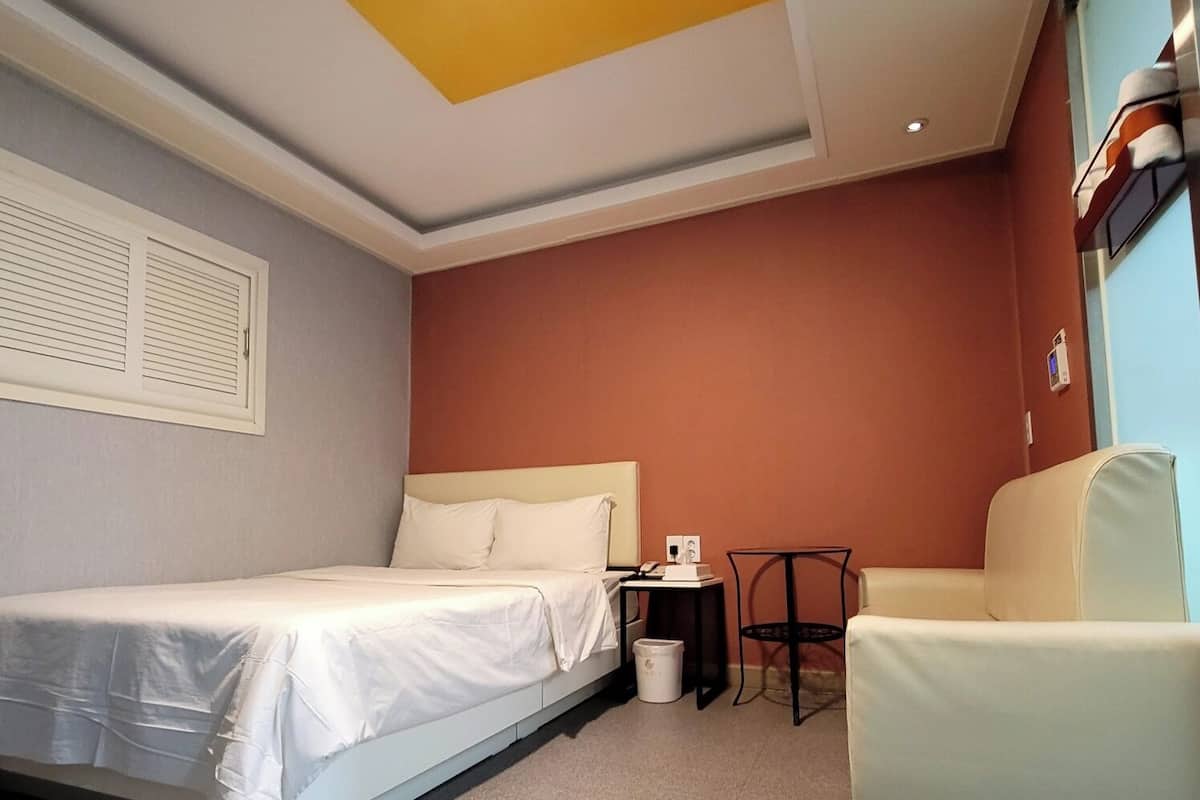 A hotel room with a bed, a chair, a small table, and a trash bin.