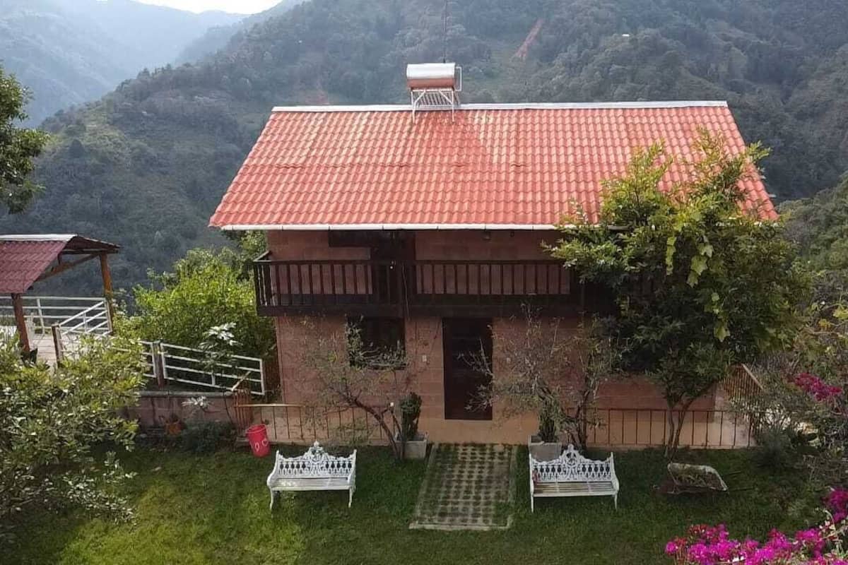 A two-story house with a red-tiled roof, surrounded by lush greenery and hills.