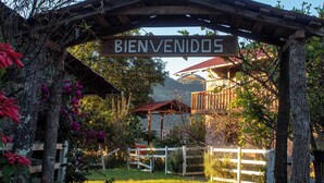 A wooden archway with a "BIENVENIDOS" sign, surrounded by lush greenery and a mountain backdrop. - Cabañas Xakali (San Joaquín)
