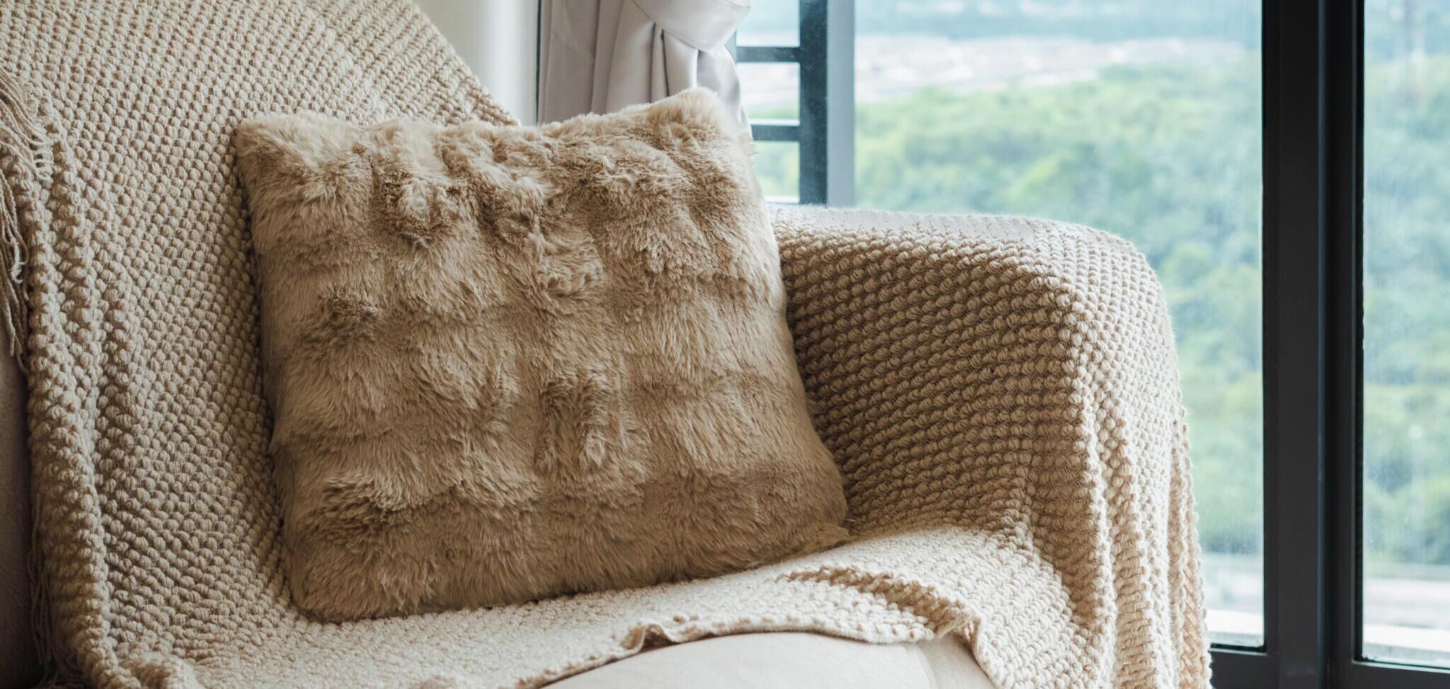 A cozy, knitted chair with a fluffy pillow, positioned near a window with a view of greenery.