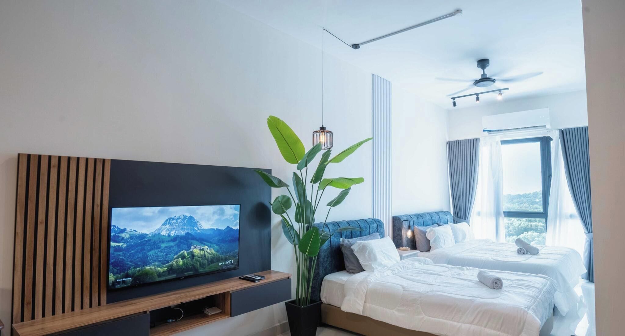 Habitación de hotel moderna con televisor de pantalla plana, una planta grande en maceta, cama con ropa blanca, ventilador de techo y ventana con cortinas.