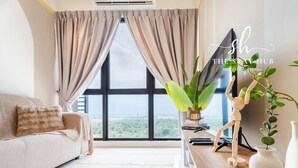 A modern living room with a large window, a sofa, a wooden desk, and a plant. - Core Soho Suites Sepang by The Stay Hub (Sepang)