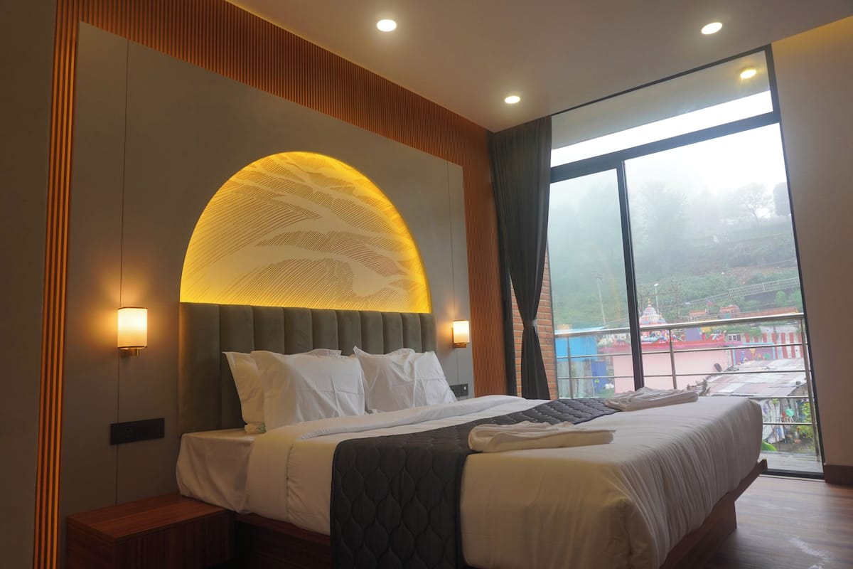 A hotel room with a large bed, a headboard with a circular light, a nightstand, and a view of a temple through a large window.
