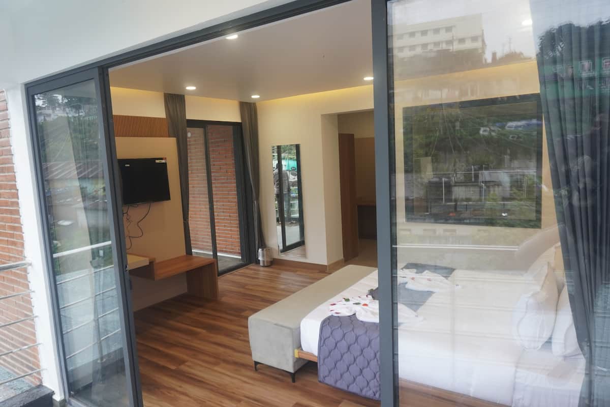 A modern hotel room with a sliding glass door leading to a balcony, a bed with a grey quilt, a small bench, and a flat-screen TV mounted on the wall.