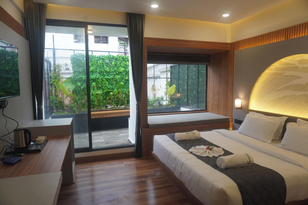 A modern hotel room with a large bed, a desk, and a view of greenery through the windows.