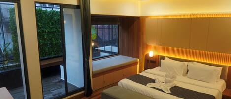 A modern hotel room with a large bed, a bench by the window, and a view of greenery outside.