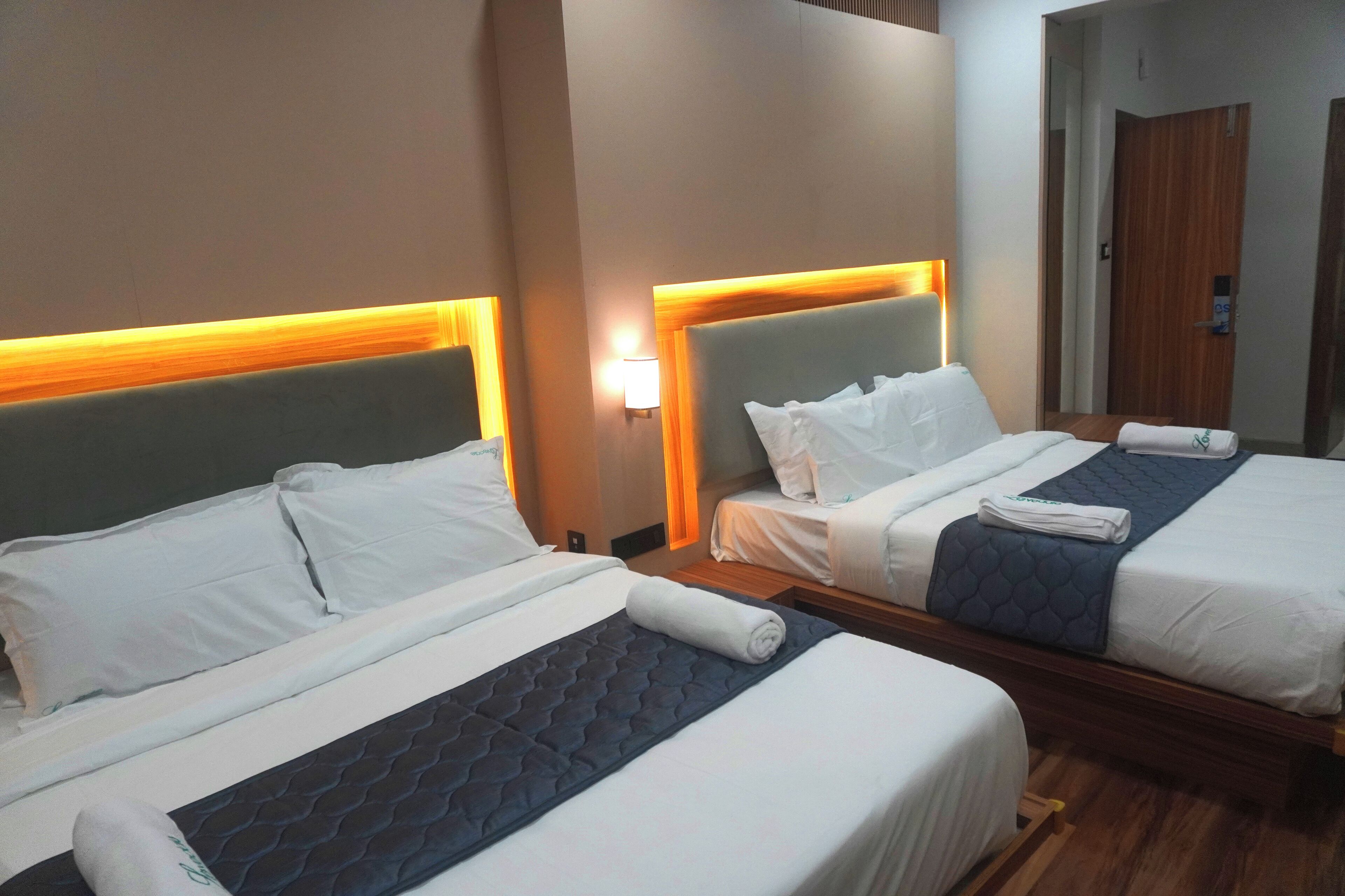 A hotel room with two beds, each with a headboard, white bedding, and a blue quilted bedspread. There is a nightstand with a lamp between the beds.