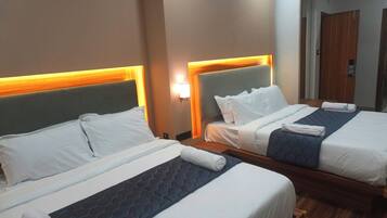 A hotel room with two beds, each with a headboard, white bedding, and a blue quilted bedspread. There is a nightstand with a lamp between the beds.