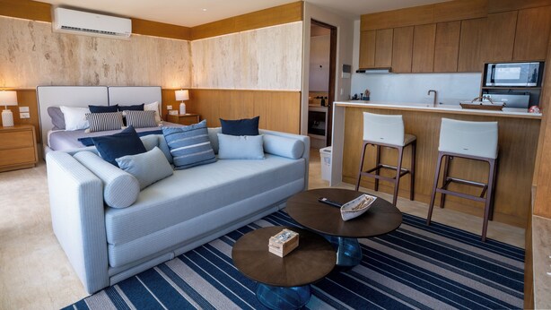 Quarto de hotel moderno com sofá, mesa de centro, área de cozinha com balcão e TV.