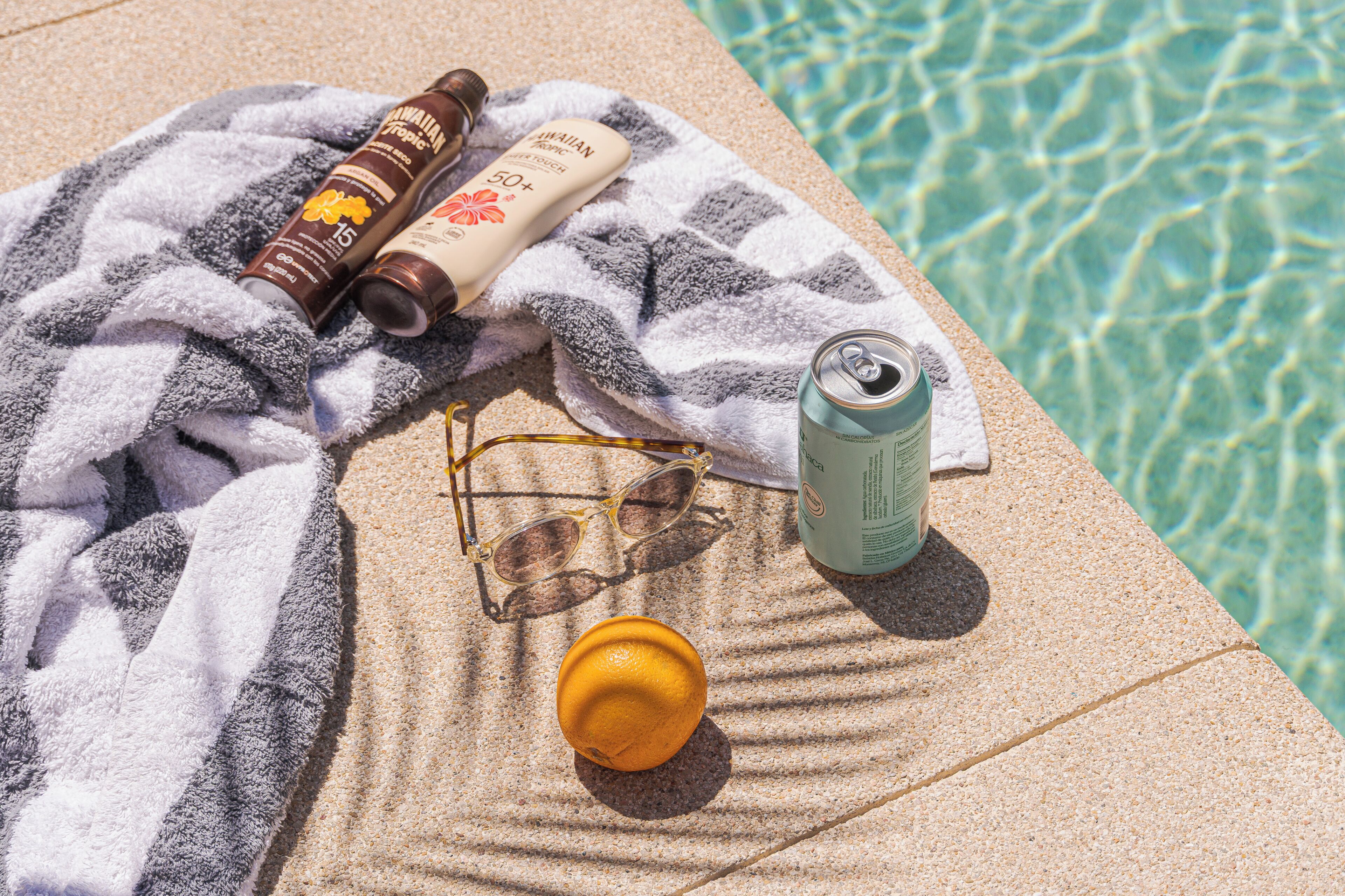 Ein Handtuch, Sonnenschutzmittel, Sonnenbrille, eine Dose und eine Orange liegen auf einer Poolumrandung.