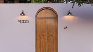 Una puerta de madera con parte superior semicircular, flanqueada por dos lámparas fijadas a la pared, y el texto 'ABASOLO CLUBHOUSE' a la izquierda.