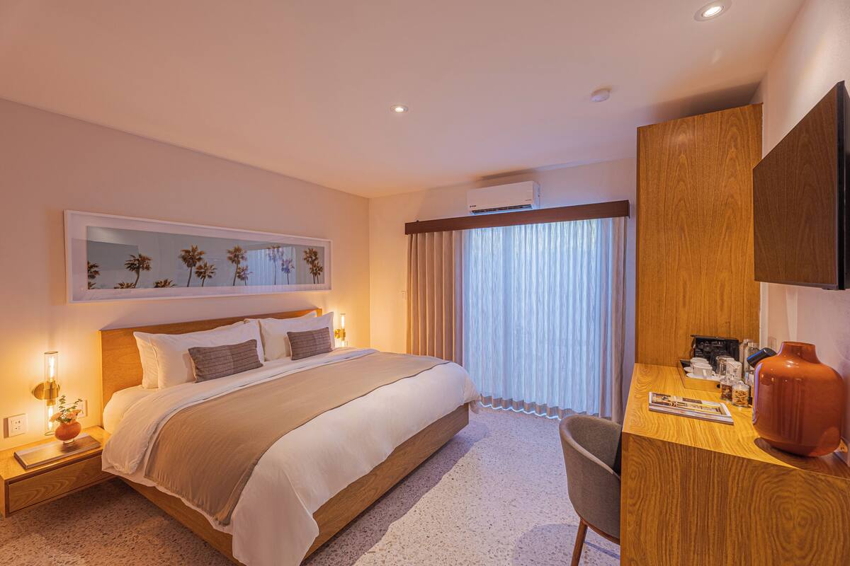 Habitación de hotel con cama, mesita de noche, escritorio, silla y un ventanal con cortinas.