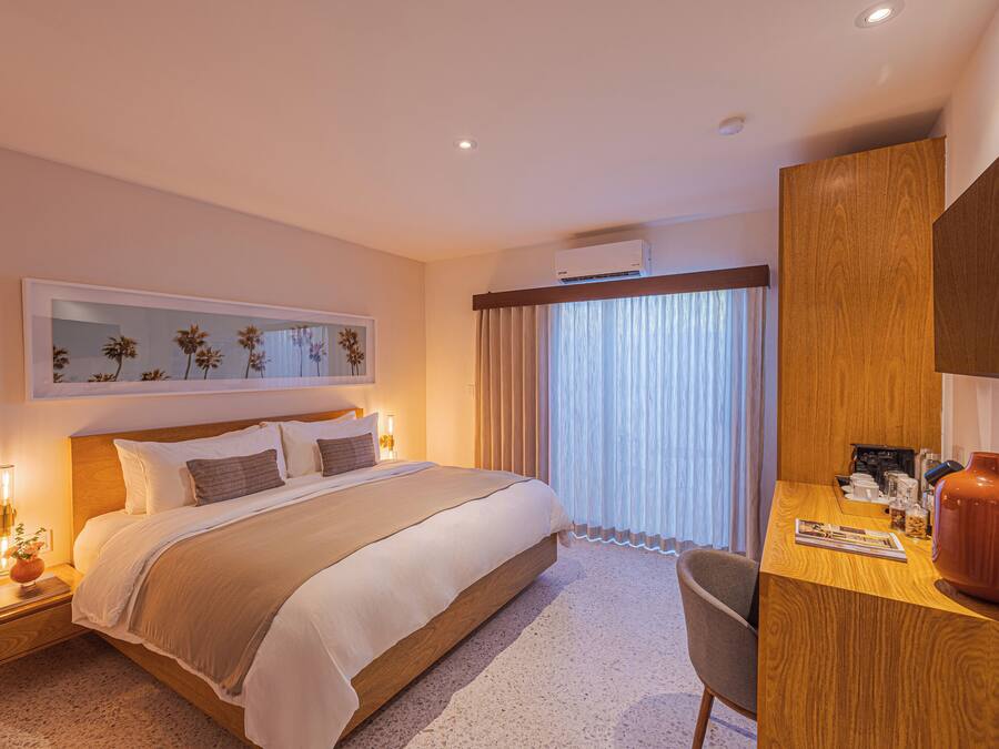 A hotel room with a bed, bedside table, desk, chair, and a large window with curtains.