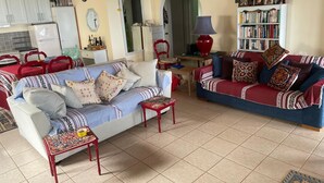 A living room with a sofa, a red armchair, a small table, and a bookshelf. There is a kitchen area with cabinets and a refrigerator in the background. - Villa Victoria (Corfu)