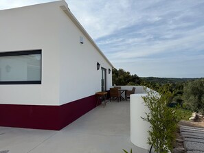 Um edifício moderno com fachada branca e base vermelha, apresentando um pátio com móveis de exterior e vista para a paisagem circundante.