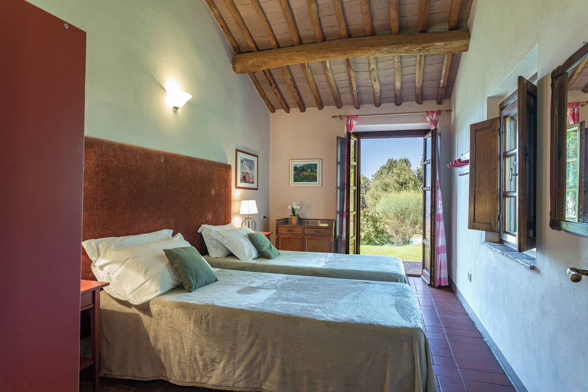 A room with two beds, a wooden ceiling, a window with curtains, and a view of greenery outside.