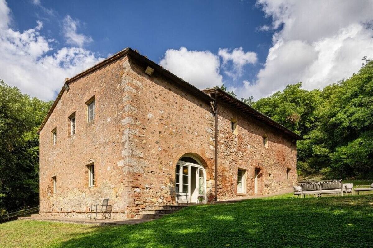 A stone building with a large arched entrance, a small porch with chairs, and a grassy area in front.