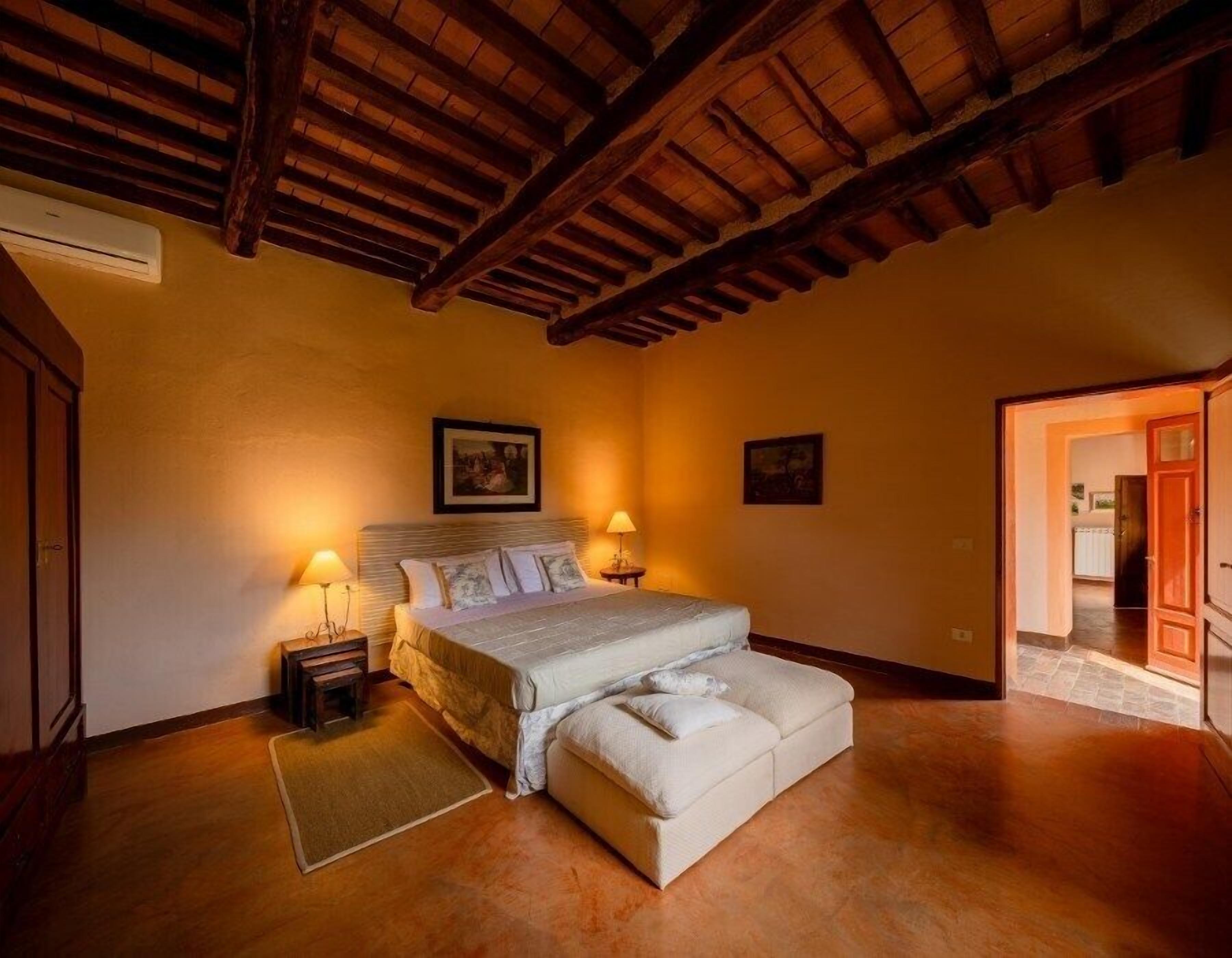 Un dormitorio con una cama grande, dos mesitas de noche con lámparas, techo de madera y vista a otra habitación por una puerta abierta.