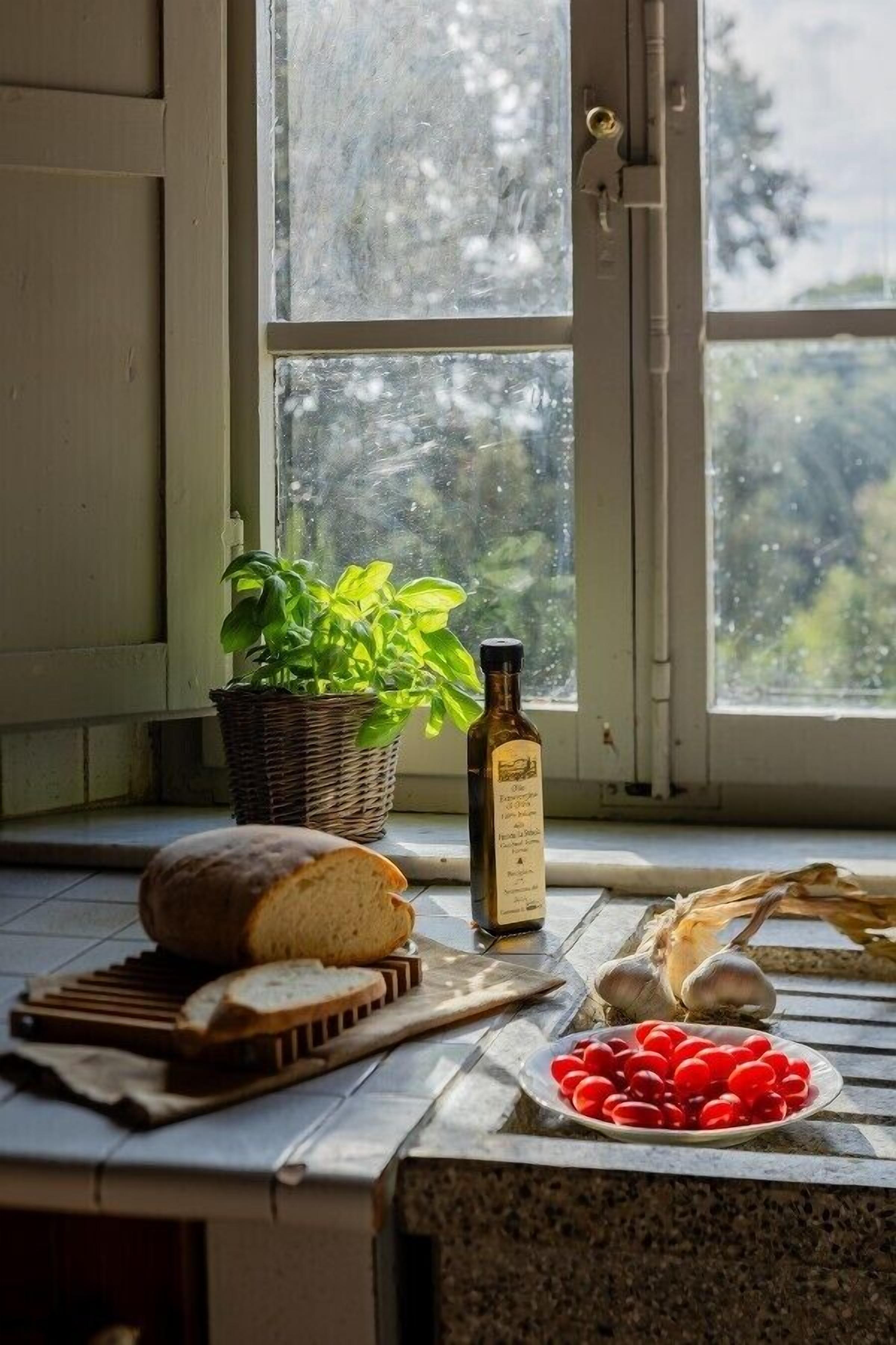 A kitchen counter with a loaf of bread, a bottle of olive oil, a basket of basil, garlic bulbs, and a plate of cherry tomatoes.