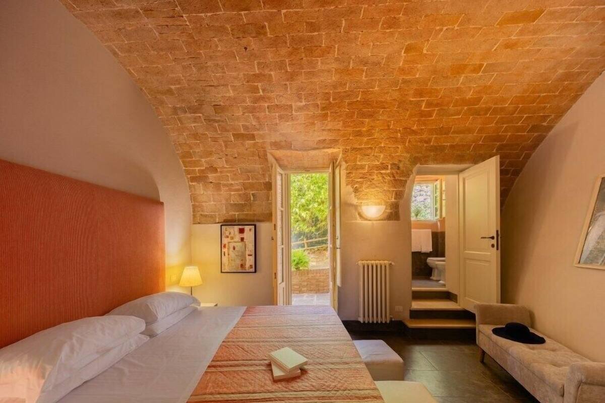 A bedroom with a brick ceiling, a bed with a striped bedspread, a bedside table with a lamp, and a view of a bathroom and outdoor area through an open door.