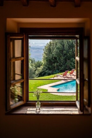 A window with an open view of a swimming pool, a grassy area, and a forested landscape. - Fattoria La Striscia (Gambassi Terme)