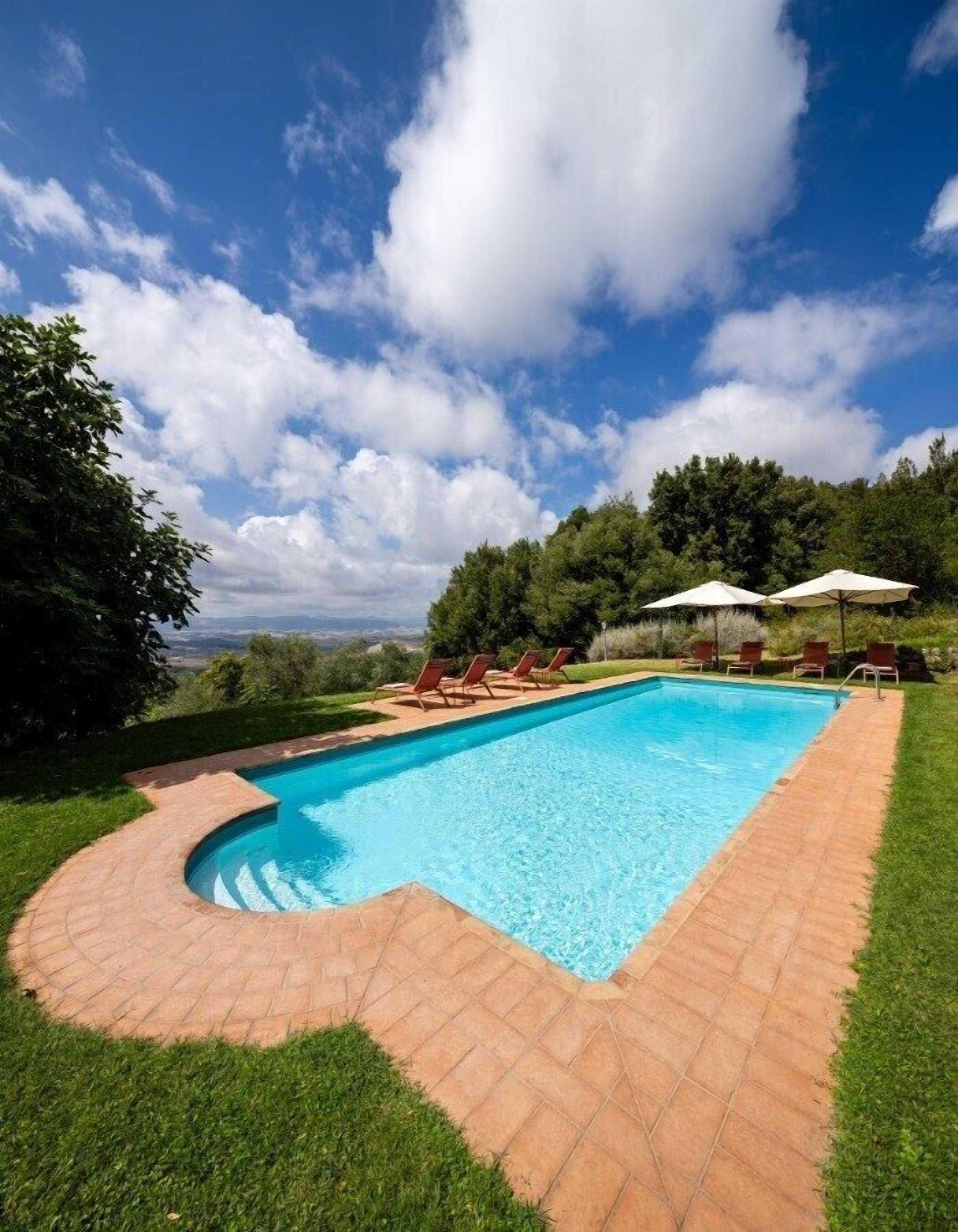 Une piscine rectangulaire à bords incurvés, entourée d’une terrasse en briques. On y trouve des chaises longues et des parasols.