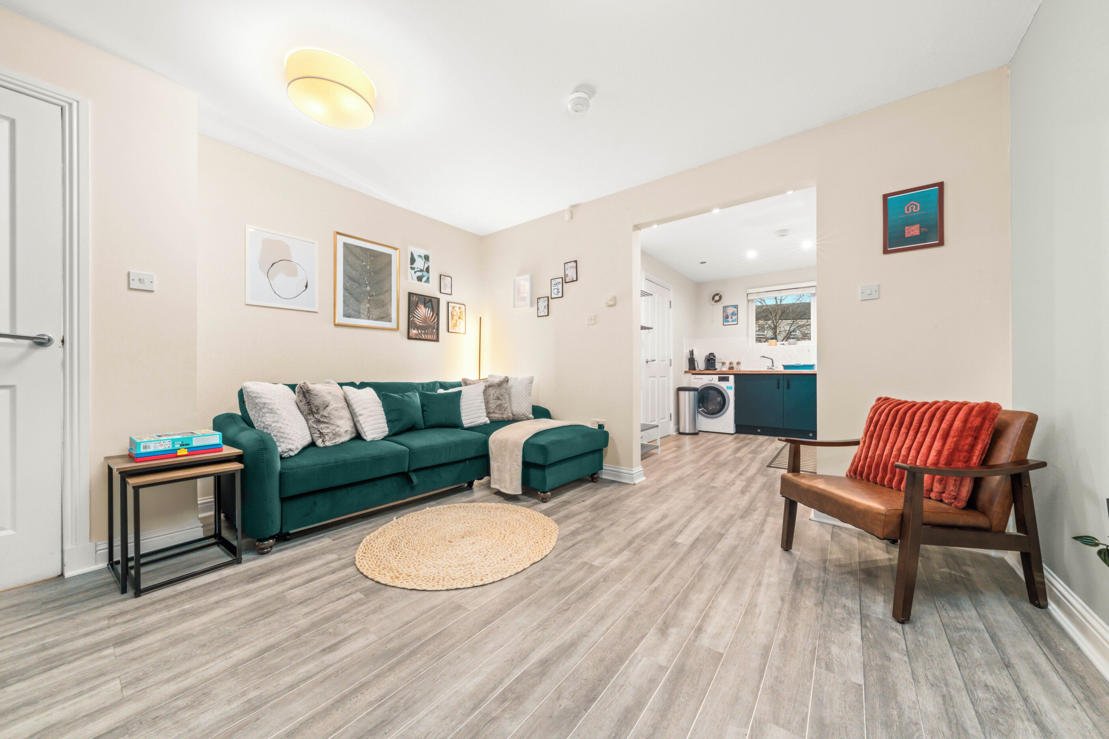 Un salon avec un canapé vert, un fauteuil en cuir marron, un tapis rond et, au fond, un coin cuisine.