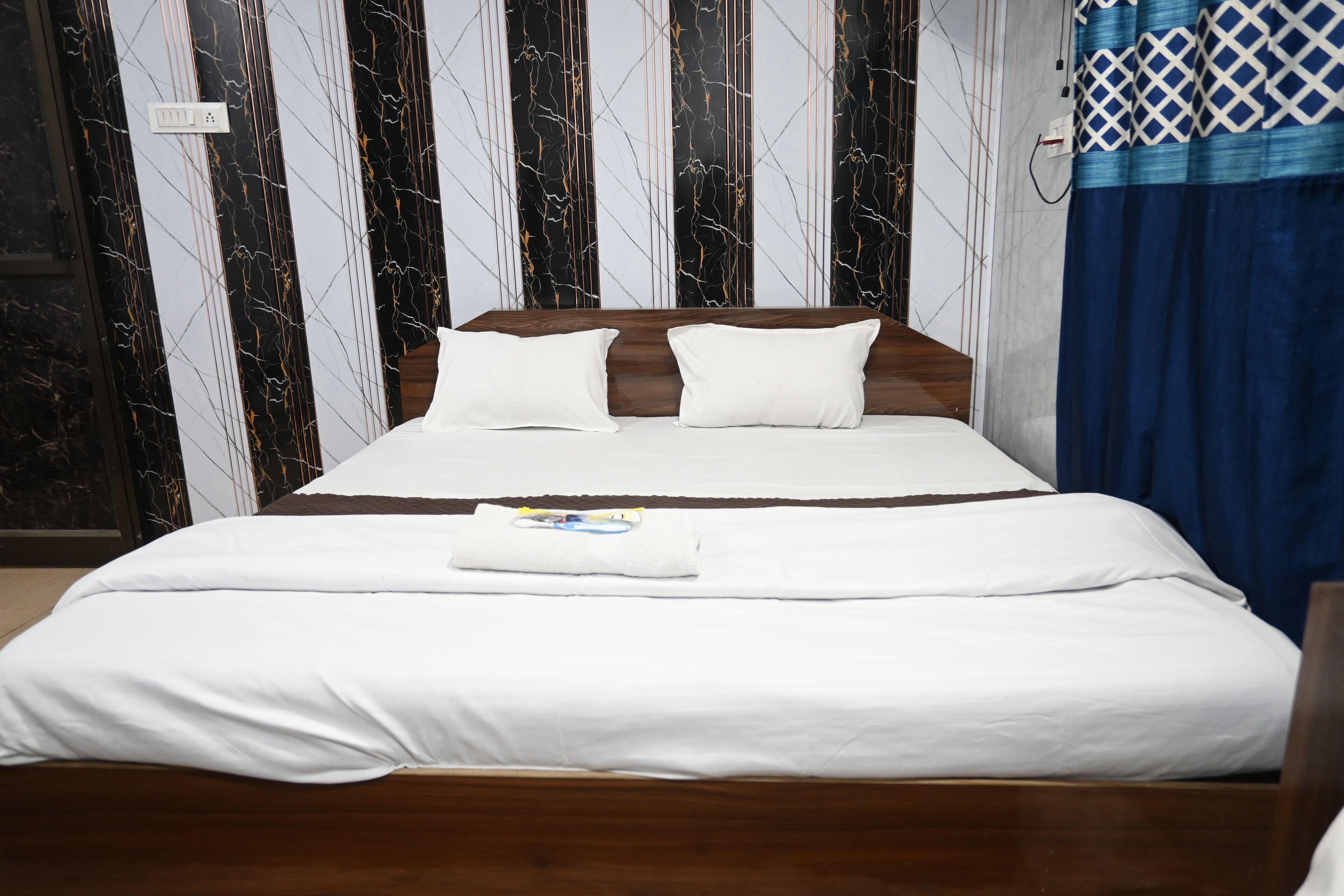 Una cama bien hecha con sábanas y almohadas blancas, un cabecero de madera y una pared con estampado de mármol al fondo.