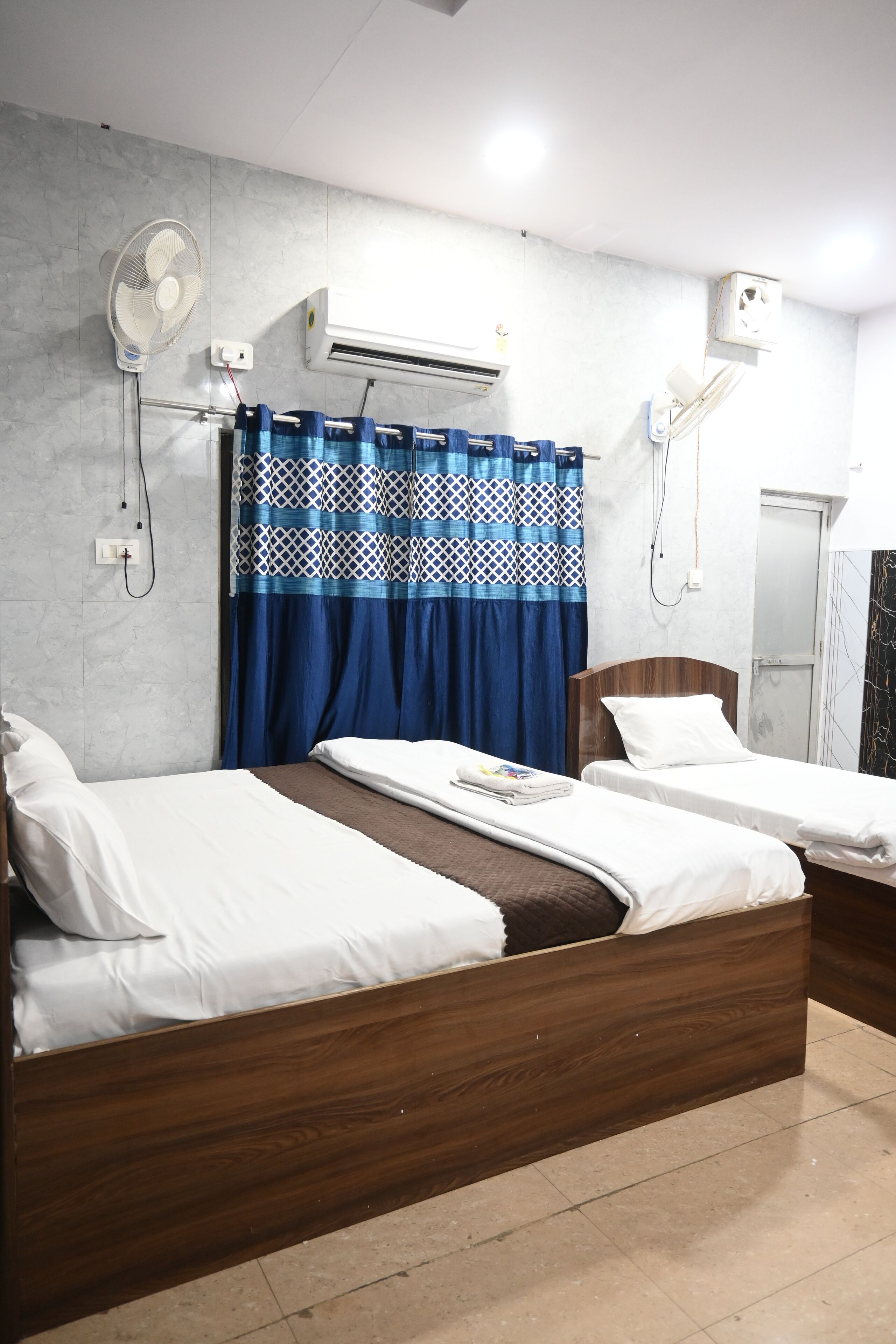 A hotel room with two beds, a large window with blue curtains, an air conditioning unit, and a wall-mounted fan.