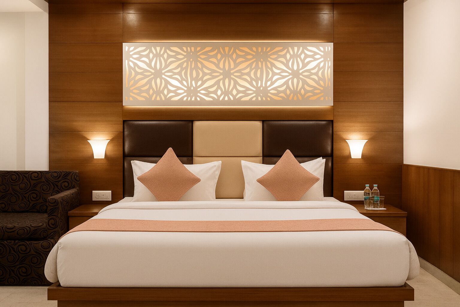 A hotel room with a large bed, two bedside lamps, a decorative wall panel, a nightstand with water bottles, and a patterned armchair.