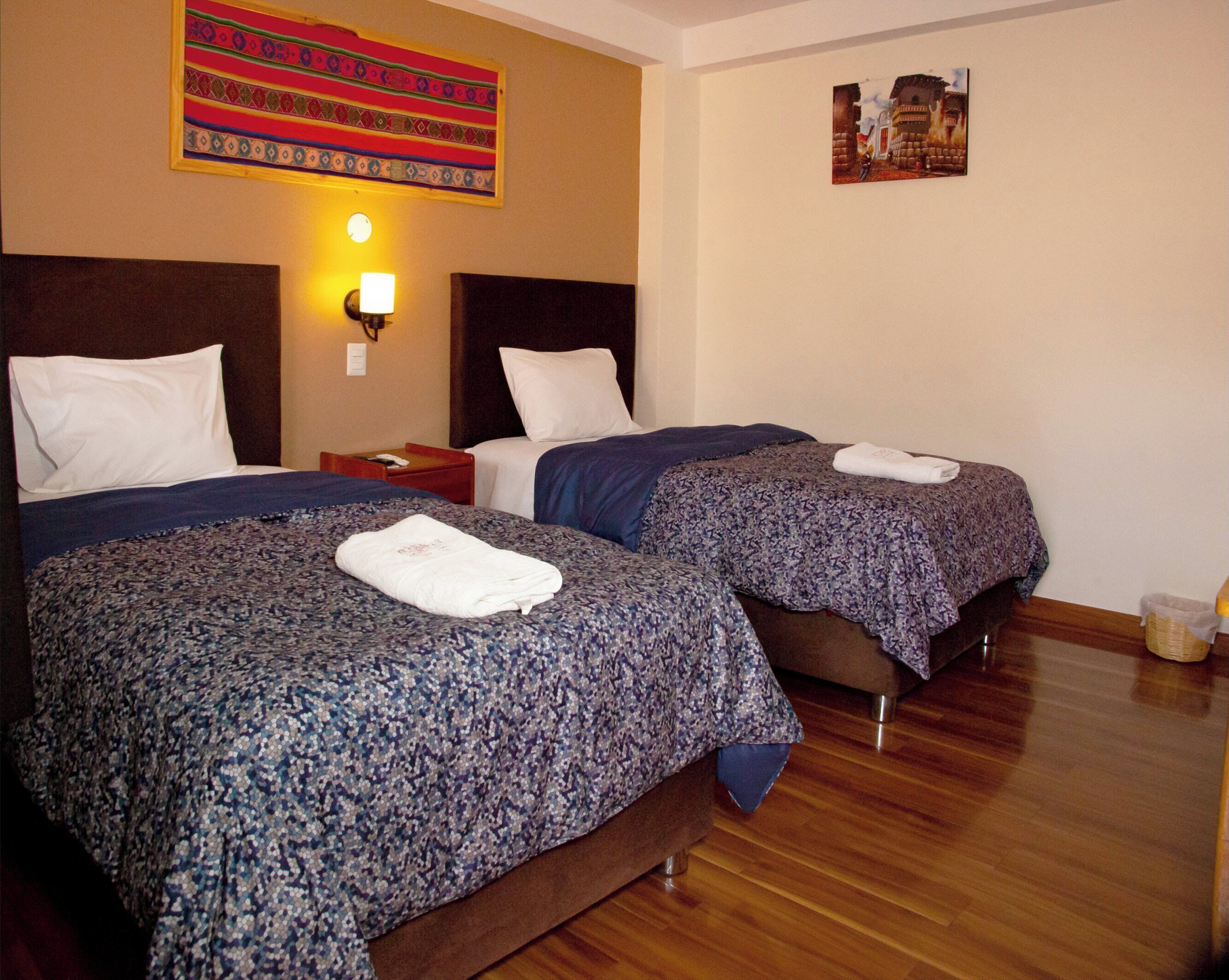 A hotel room with two beds, each with a patterned bedspread and a folded towel on top. There is a nightstand with a lamp between the beds, and a wall hanging above the beds.