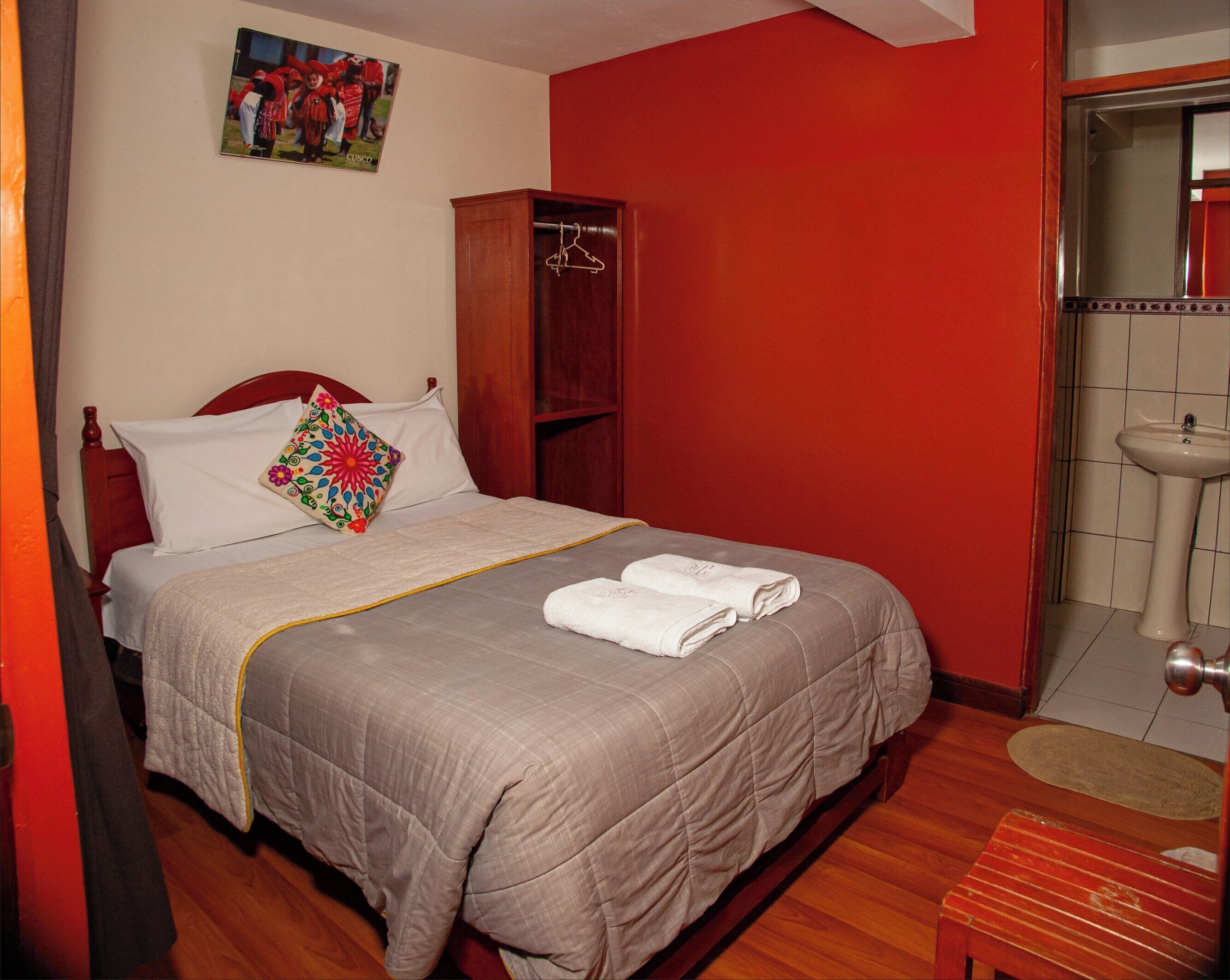 A neatly made bed with a grey quilt, white pillows, and a colorful pillow. There is a wooden wardrobe, a red wall, and a bathroom with a sink and mirror.