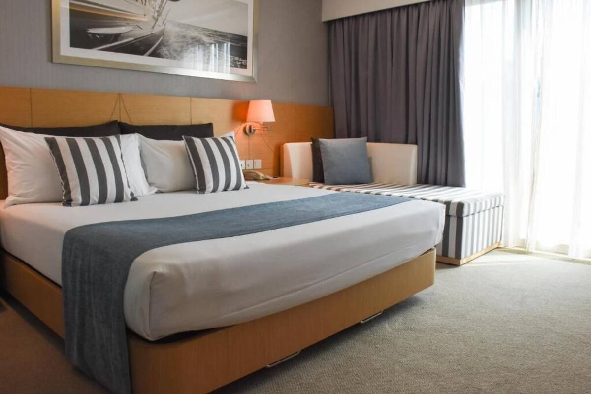 A hotel room with a large bed, a bedside table with a lamp, a window with curtains, and a framed picture on the wall.