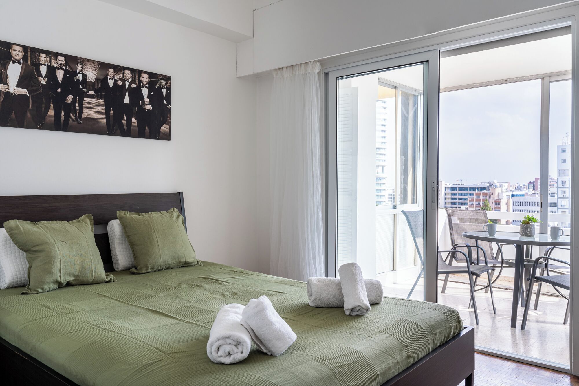 A hotel room with a bed, pillows, a green bedspread, a balcony with a table and chairs, and a city view.