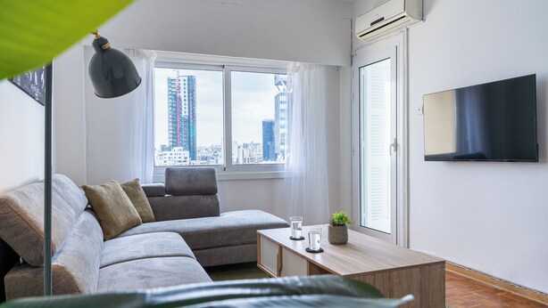 A modern living room with a sofa, a coffee table, a TV, and a view of the cityscape through large windows. - Bastia - Skyline 1-BR Apt in Nicosia (Nicosia)