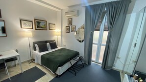 A hotel room with a bed, desk, and chair, a small table with items, and a view through a window with curtains. - Short Rent Milano Porta Vittoria (Milan)