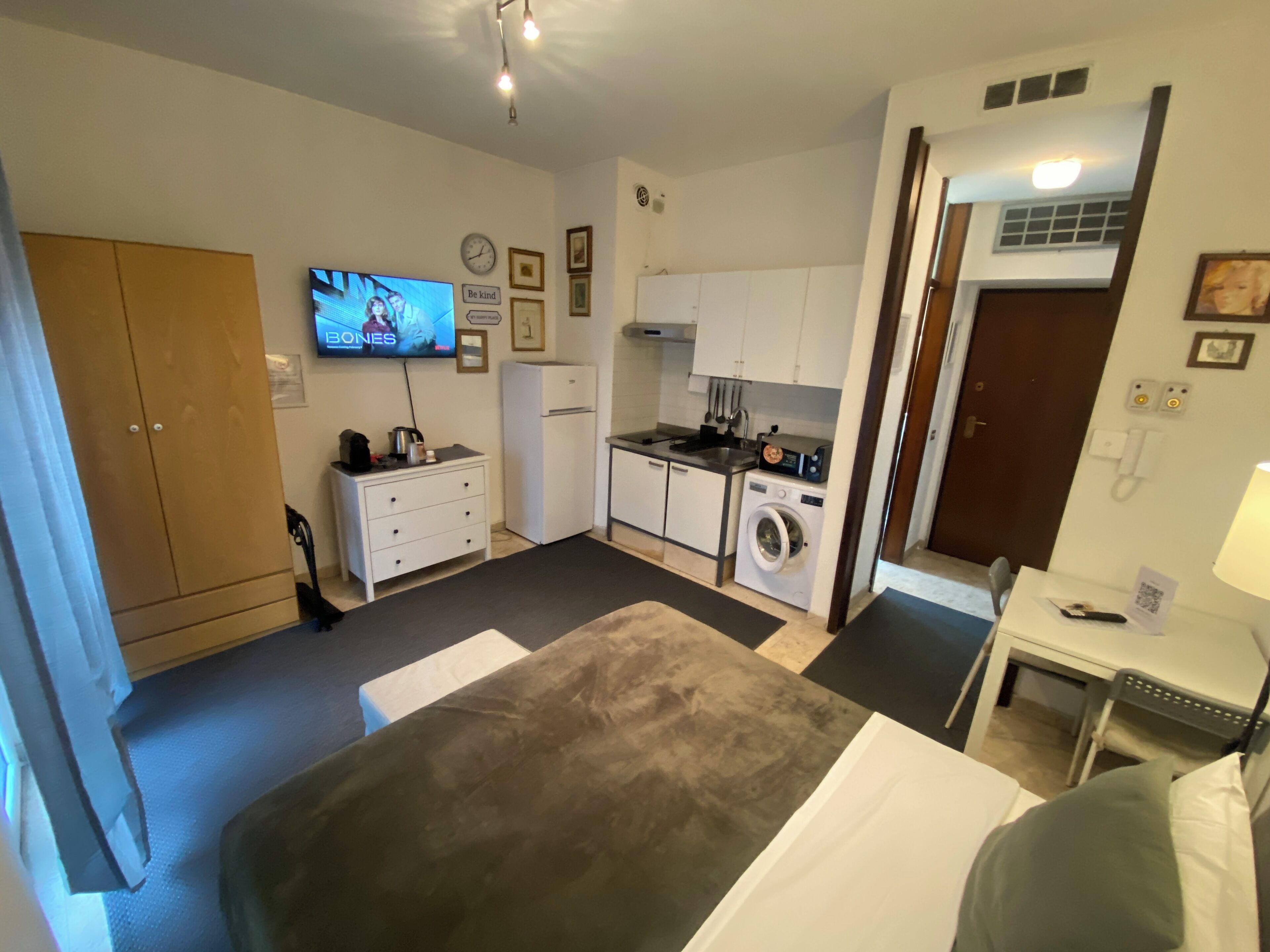 A compact hotel room with a bed, a small kitchenette, a washing machine, a TV, and a wardrobe.