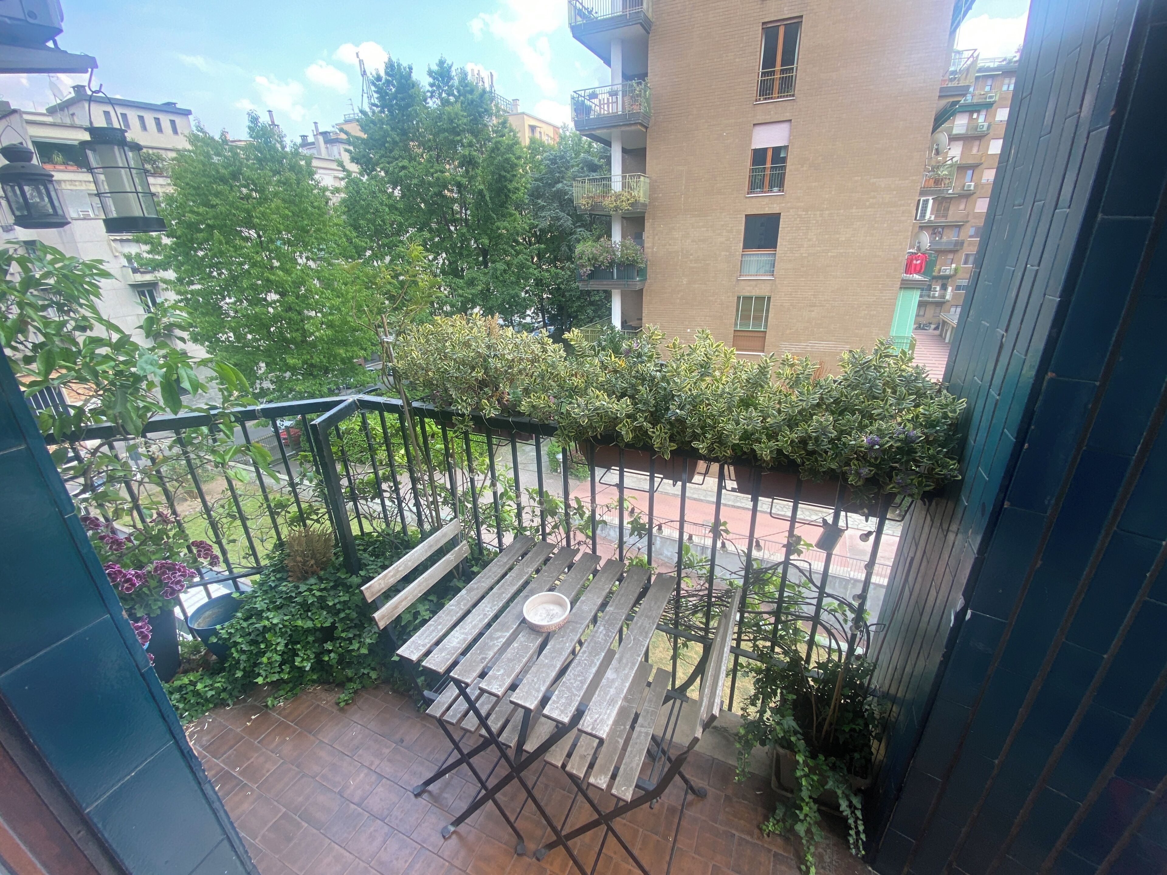 A balcony with a wooden table and chairs, surrounded by greenery and a view of a multi-story building.