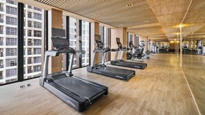 A gym with treadmills aligned along a wall with large windows, wooden flooring, and a modern ceiling design. - Minori by Kozystay - Alam Sutera (South Tangerang)