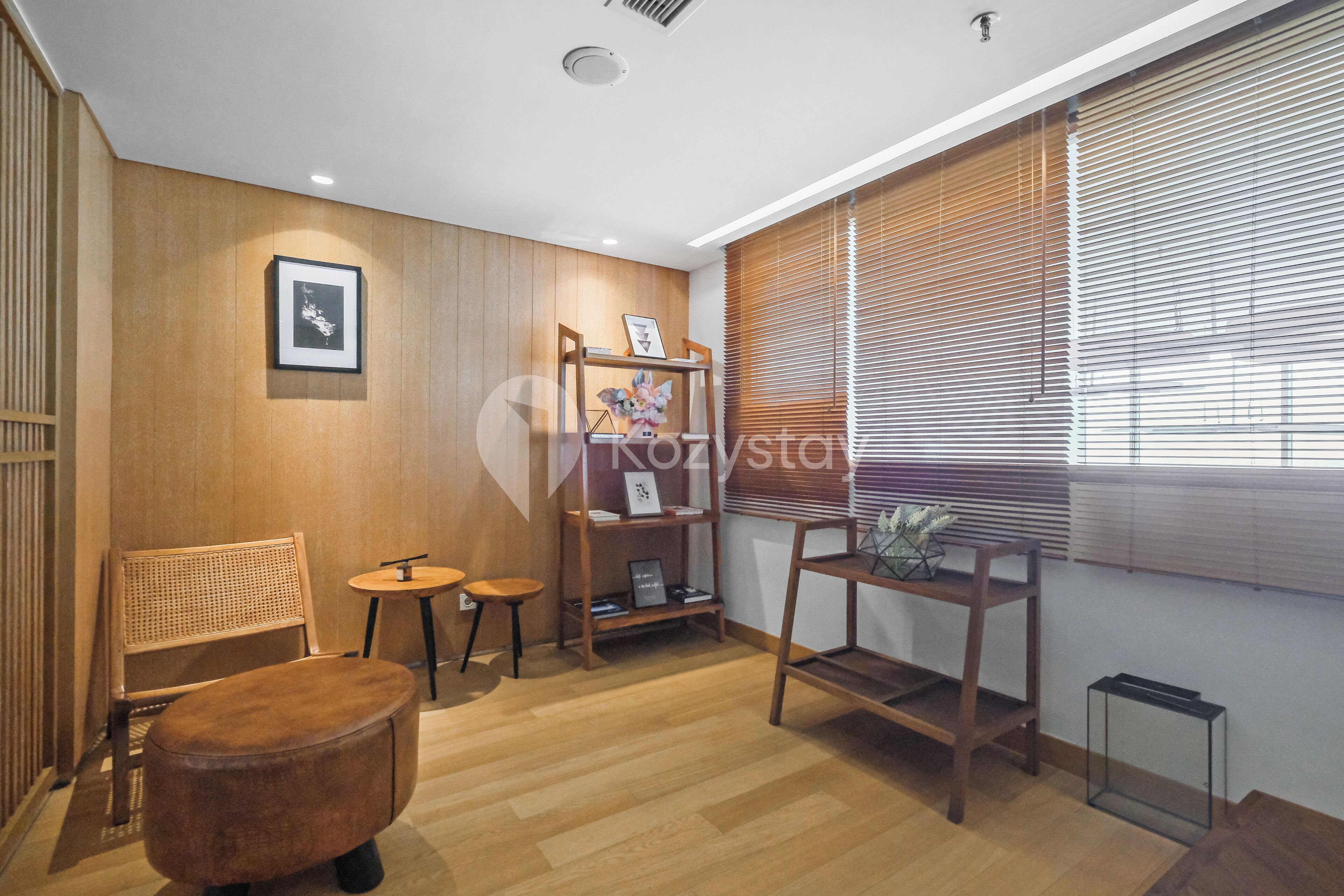 Una habitación moderna con suelo de madera, una mesa redonda de madera, un banco acolchado, una estantería y una ventana con persianas.
