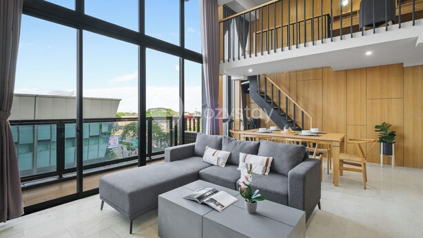 A modern living room with a grey sectional sofa, a coffee table, and a dining area with a wooden table and chairs. A staircase leads to an upper level. - Minori by Kozystay - Alam Sutera (South Tangerang)