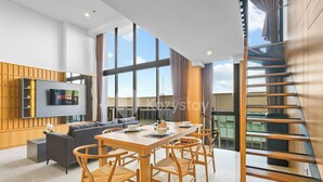 A modern living room with a large TV, a dining area with wooden furniture, and a view of the sky through large windows. - Minori by Kozystay - Alam Sutera (South Tangerang)
