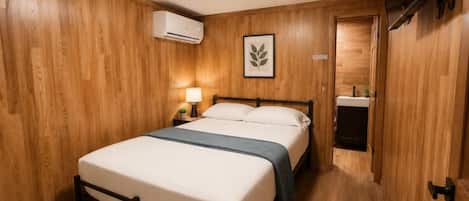 A single bed with a headboard, a bedside table with a lamp, an air conditioning unit, and a television mounted on the wall.