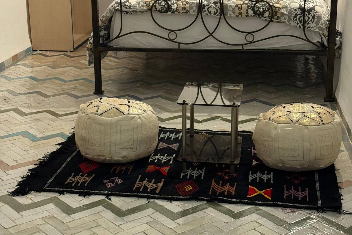 Une chambre à coucher avec un lit en métal, une literie à motifs, deux coussins rouges, une armoire et deux tabourets décoratifs posés sur un tapis à motifs.