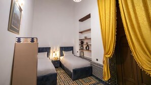 A room with two beds, a nightstand, a lamp, a framed picture, and a shelf with decorative items. - dar fes flower (Fès)