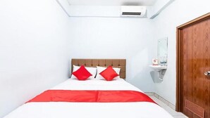 A hotel room with a bed, a wooden door, a white wall, and a small white shelf with toiletries. - Hotel Cemara Gading (Dramaga)