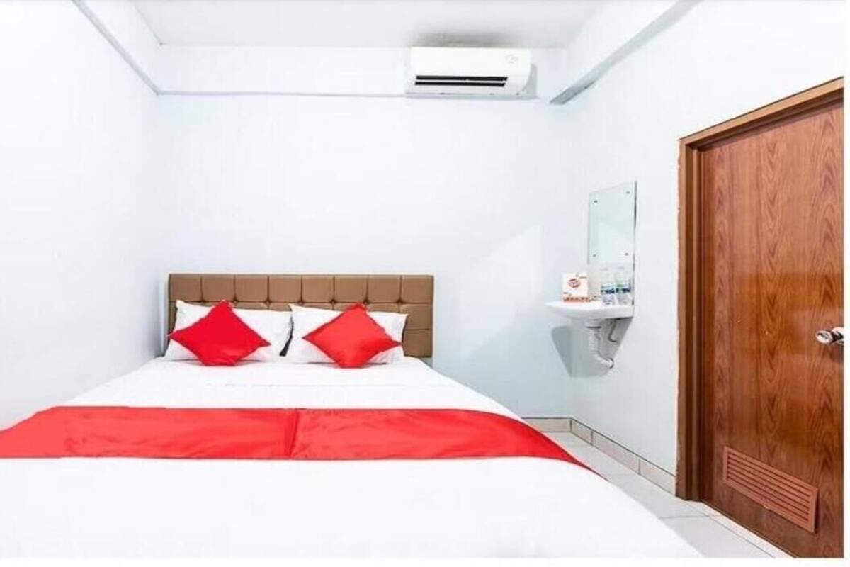 A bedroom with a bed, a wooden door, a white wall, and a small shelf with toiletries.